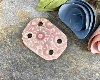 Op de afbeelding: Een roze keramische knoop met een bloemenpatroon en vier gaten. De knoop ligt op een grijs, getextureerd oppervlak.