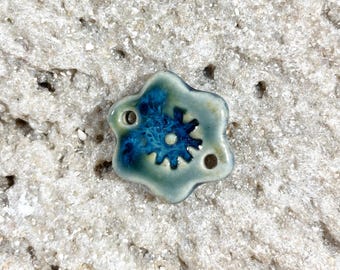 May include: A ceramic bead with a six-petal flower shape. The bead has a light blue-green base color with a dark blue flower design in the center. Two small holes are visible for stringing.