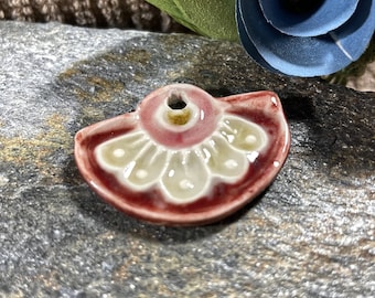 May include: A ceramic pendant in the shape of a half-circle with a floral design. The pendant is red with white and yellow accents.