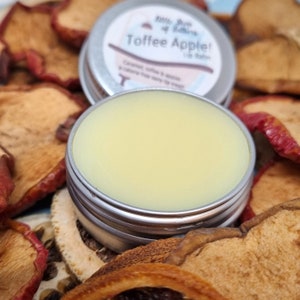 May include: A silver tin of Toffee Apple lip balm, with a light yellow balm visible. The lid of a second tin is behind it. The lid reads "Toffee Apple! Lip Balm". Dried apple slices are scattered around the tins.
