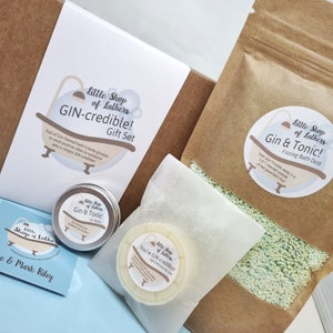 May include: A gift set with a gin theme, including a bath bomb, lip balm, and a solid moisturiser. The bath bomb is in a brown paper bag with a white label that says "Little Shop of Lathers Gin & Tonic! Fizzing Bath Dust". The lip balm is in a small tin with a white label that says "Little Shop of Lathers Gin & Tonic Lip Balm". The solid moisturiser is in a white paper bag with a white label that says "Little Shop of Lathers You're GIN-credible! Solid Moisturiser Bar".