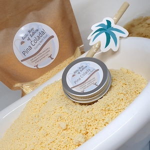 May include: A white bath tub filled with yellow bath salts. A brown paper bag with the text "Little Shop of Lathers Pina Colada! Fizzing Bath Dust" is visible in the background. A small silver tin with the text "Little Shop of Lathers Pina Colada Lip Balm" is sitting on top of the bath salts. The tin has a tropical pineapple and coconut design.