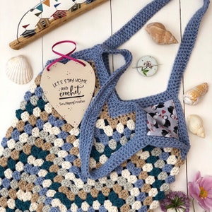 May include: A blue crocheted tote bag with a brown, white, and teal granny square pattern. The bag has a wooden heart-shaped sign that says "Let's Stay Home and Crochet" with the words "SewHappyCreative" at the bottom.