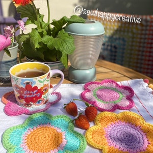 May include: Three crocheted flower-shaped coasters in pink, yellow, and green with a light blue mug with the text "Keep Growing" on a white surface with strawberries.