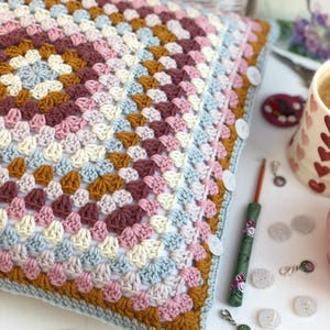 May include: A square crochet pillow cover featuring a multi-colored granny square pattern in shades of dusty pink, mustard yellow, cream, and light blue. The edge is finished with white buttons, and a green floral crochet hook lies nearby.