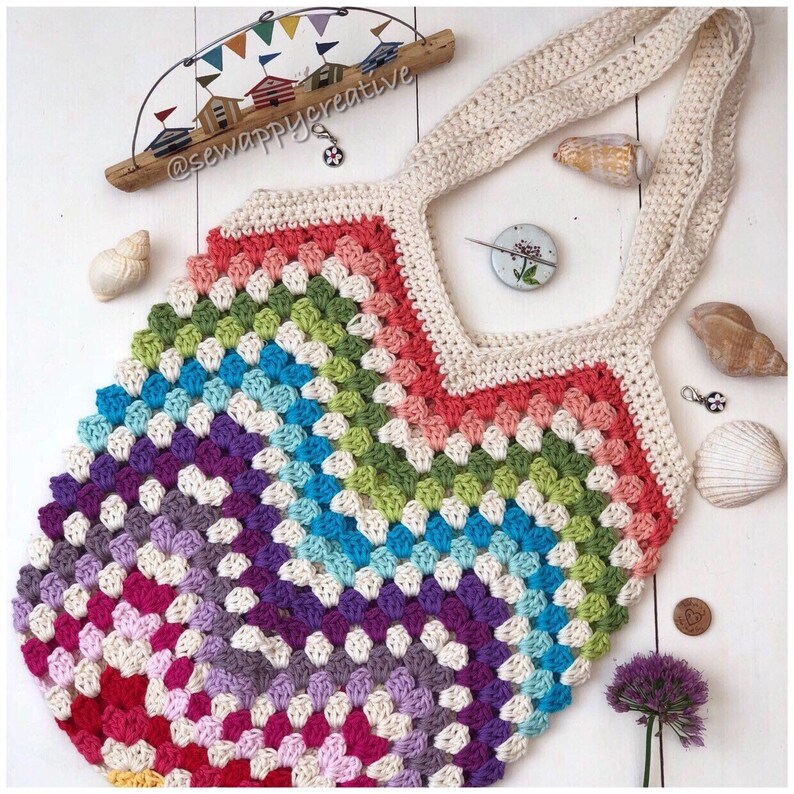 May include: A colorful crocheted tote bag with a rainbow pattern and cream-colored straps. The bag is made with a granny square stitch and has a unique, handmade look.