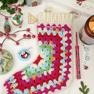 Può includere: Una calza di Natale fatta a mano, all'uncinetto con un motivo a granny square. La calza presenta un polsino color crema e un corpo di quadrati vibranti in rosso, rosa, blu e verde. Il testo "@sew happy creative" è visibile.