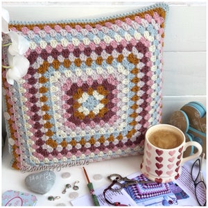 May include: A square crochet pillow cover featuring a multi-colored granny square pattern in shades of light blue, pink, cream, and mustard yellow. The cover has white buttons along the side edge and is displayed with a heart-patterned mug.