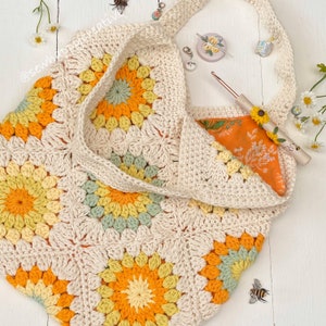Puede incluir: Una bolsa de mano de crochet con un patr&oacute;n floral en tonos amarillos, naranjas y verdes. La bolsa tiene una correa larga y est&aacute; hecha con hilo blanco.