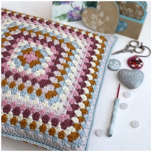 May include: A square crochet pillow cover featuring a multi-colored granny square pattern in shades of dusty pink, burgundy, mustard yellow, and light blue. A crochet hook with the text @sewhappycreative is placed next to the pillow.