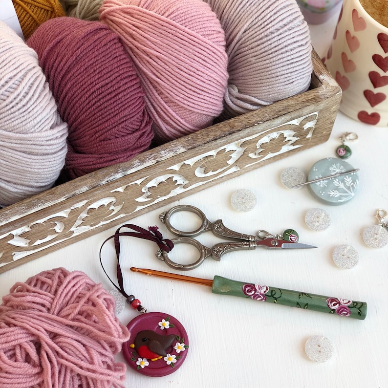 May include: A wooden tray filled with balls of yarn in shades of pink and mauve, accompanied by metal embroidery scissors, a green crochet hook with floral patterns, a round robin pendant, and several small white buttons.