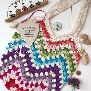 May include: A colorful crochet tote bag with a rainbow gradient pattern. The bag is made with a variety of colors, including red, orange, yellow, green, blue, indigo, and violet. The bag has a heart-shaped wooden sign hanging from it that says "Let's Stay Home and Crochet".