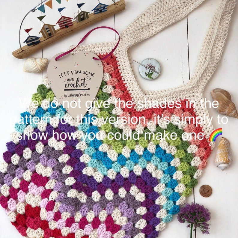 May include: A colorful crochet tote bag with a rainbow gradient pattern. The bag is made with a variety of colors, including red, orange, yellow, green, blue, indigo, and violet. The bag has a heart-shaped wooden sign hanging from it that says "Let's Stay Home and Crochet".