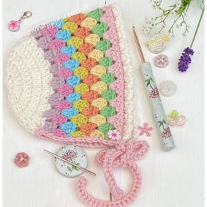 May include: A crocheted baby bonnet with a rainbow pattern and a pink tie. The bonnet is made with a variety of colors, including pink, blue, green, yellow, and white. The bonnet is tied with a pink ribbon.