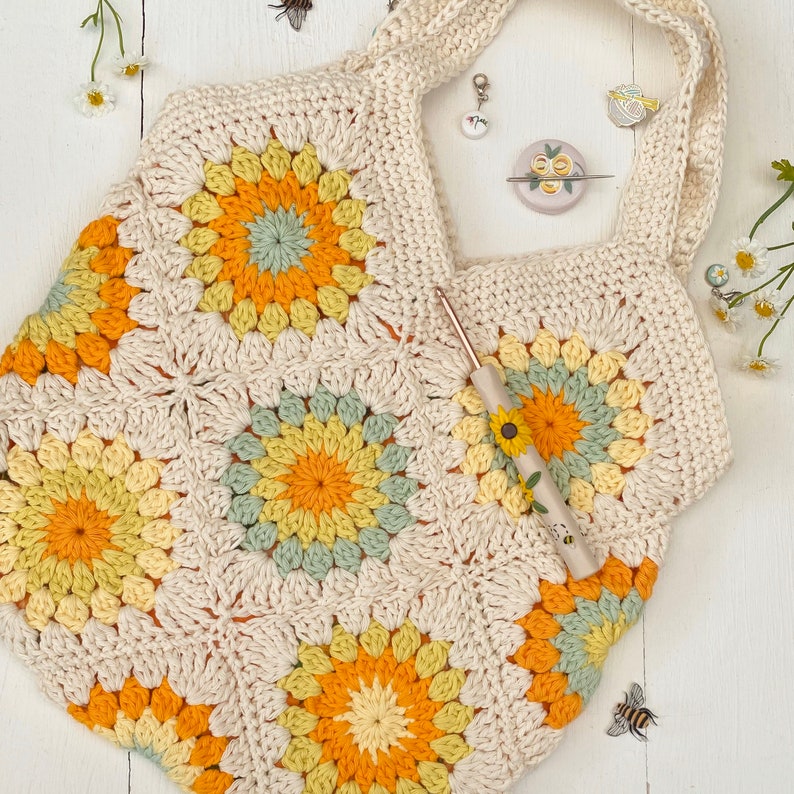 Puede incluir: Una bolsa de mano de crochet con fondo blanco y un patr&oacute;n floral amarillo, verde y naranja. La bolsa tiene una correa ancha y est&aacute; hecha con un punto de granny square.