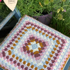 May include: A square crochet cushion cover featuring a multi-colored granny square pattern in shades of dusty pink, mustard yellow, light blue, and cream. A heart-shaped wooden sign hangs above, printed with the text Let's stay home and crochet SewHappyCreative.