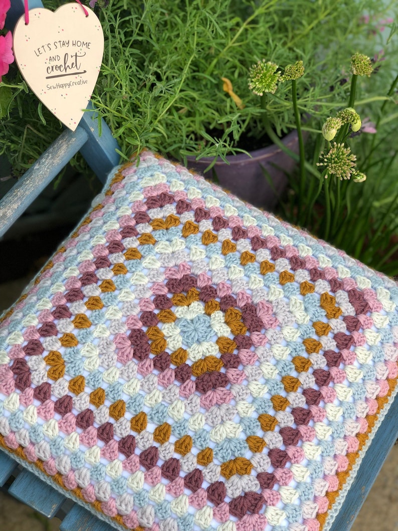 May include: A square crochet cushion cover featuring a multi-colored granny square pattern in shades of dusty pink, mustard yellow, light blue, and cream. A heart-shaped wooden sign hangs above, printed with the text Let's stay home and crochet SewHappyCreative.