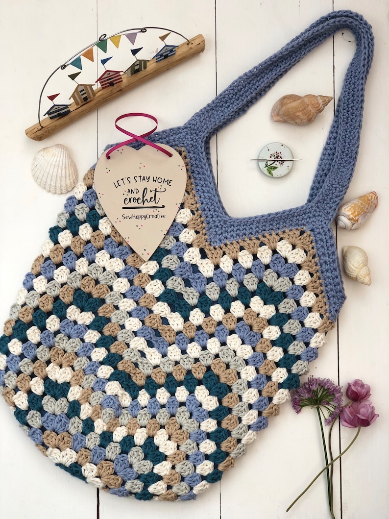 May include: A crocheted tote bag with a blue, beige, white, and teal color pattern. The bag has a blue handle and a wooden heart-shaped tag that says "Let's stay home and crochet".