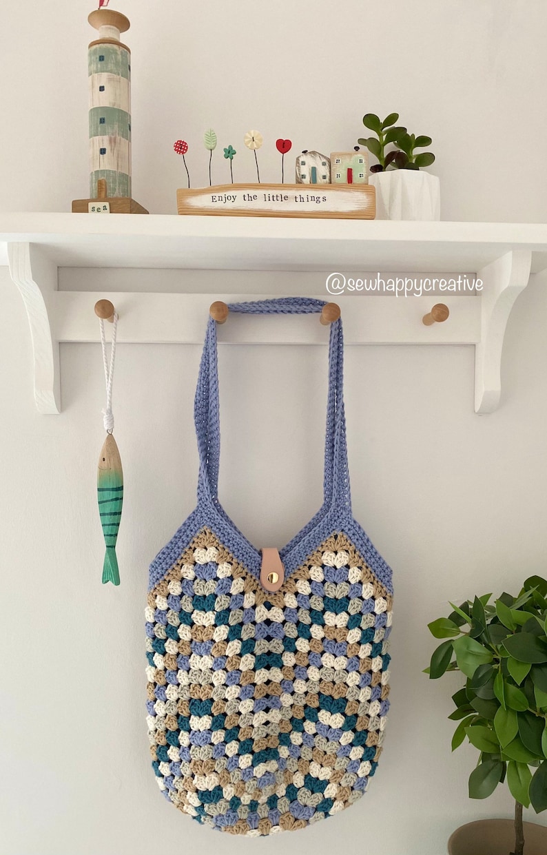 May include: A crocheted tote bag with blue, beige, white, and green granny squares. The bag has a light blue strap and a brown leather handle. The bag is hanging on a white shelf with a wooden fish and a wooden lighthouse figurine.