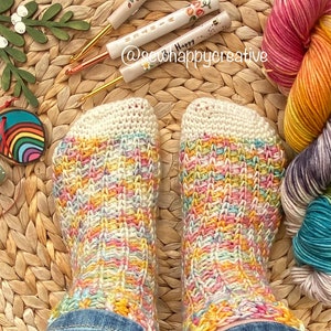 May include: A pair of colourful crochet socks made with a multi-coloured yarn. The socks are on a woven straw surface with crochet hooks and balls of yarn nearby. The text "@sewhappycreative" is visible in the image.