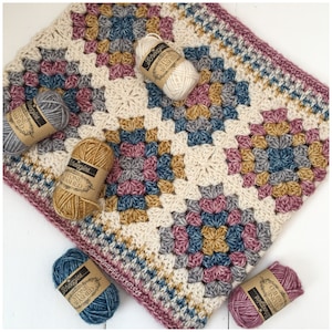 Op de afbeelding: Een gehaakt deken met een granny square patroon in tinten crème, roze, blauw en goud. Het deken is omringd door bollen garen in dezelfde kleuren, elk met het label "Scheepjes Stone Washed".