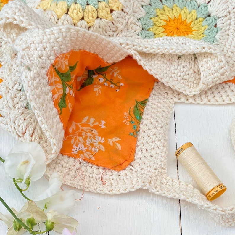 Puede incluir: Una bolsa de mano de crochet con un forro de tela floral. La bolsa est&aacute; hecha con hilo blanco y tiene un dise&ntilde;o de granny square. Un carrete de hilo blanco es visible en el fondo.