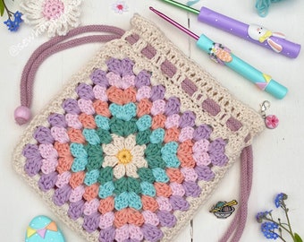 Op de afbeelding: Een gehaakte trekkoordtas met een bloemenpatroon in tinten roze, groen en blauw. De tas is gemaakt met een granny square-ontwerp en heeft een witte achtergrond. De tas ligt op een wit oppervlak met een paar kleine bloemen en haaknaalden.