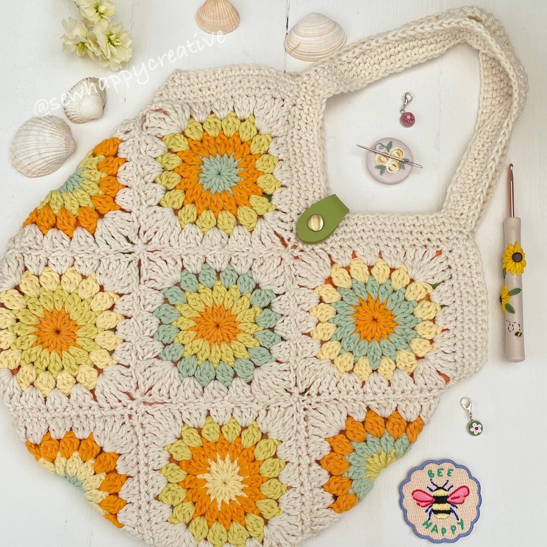 Puede incluir: Un bolso de mano de ganchillo de forma granny square con una correa beige y detalles verdes. El bolso tiene un patr&oacute;n floral con colores amarillo, naranja y verde. El bolso est&aacute; sobre una superficie blanca con otros art&iacute;culos de ganchillo y flores.