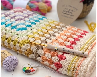 Puede incluir: Una manta de ganchillo colorida con un patrón de arcoíris en tonos de rosa, naranja, amarillo, verde, azul y morado. La manta está hecha con un punto de granny square y tiene un aspecto texturizado y esponjoso. También se muestra un gancho de ganchillo y un pequeño marcador de punto de cerámica con temática de abejas.