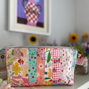 May include: A rectangular zippered pouch constructed from vertical fabric strips featuring various floral, strawberry, and gingham patterns in shades of pink, yellow, green, and white. A small pink tag with the text Good Vibes Always is attached.