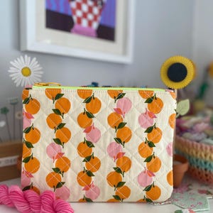 May include: A quilted pouch with a repeating pattern of orange and pink citrus fruits on a cream background. The pouch has a neon green zipper and is displayed with pink yarn and decorative flowers.