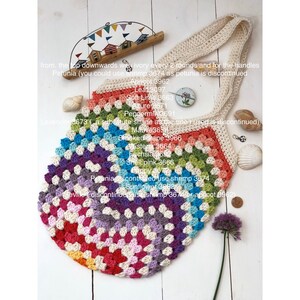 May include: A colorful crocheted tote bag with a rainbow pattern. The bag has a white handle and is made with a variety of different colored yarn. The bag is sitting on a wooden surface with a few seashells and a small bouquet of purple flowers.