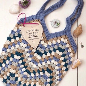 May include: A crocheted tote bag with blue, beige, white, and teal granny squares. The bag has a blue handle and a wooden heart-shaped tag that says "Let's stay home and crochet".