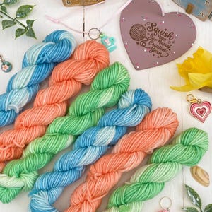 May include: Vibrant yarn skeins in blue, coral, and green are displayed. A heart-shaped sign with the text "Squish Your Sew Happy Creative Yarn" is visible. Decorative charms and miniature houses are also present, suggesting a crafting theme. The yarn is arranged on a white surface.