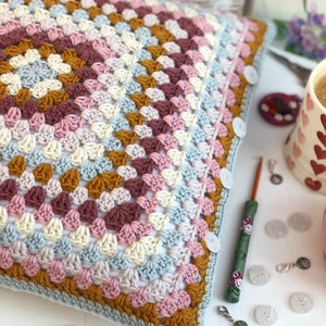 May include: A square crochet pillow cover featuring a granny square pattern in shades of dusty pink, mustard yellow, cream, and light blue. The edge is finished with a light blue border and white buttons. A green floral crochet hook lies nearby.
