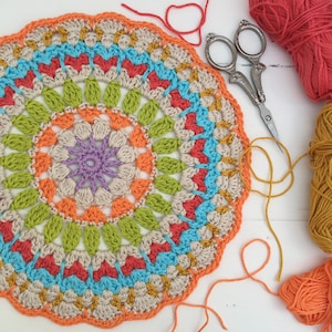 May include: A colorful crocheted mandala with orange, red, green, blue, and purple yarn. The mandala is on a white surface with yarn and scissors.