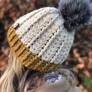 May include: A white and mustard yellow crocheted beanie with a gray faux fur pom pom. The beanie has a button on the side.