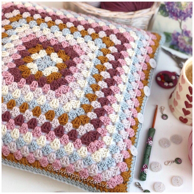 May include: A square crochet pillow cover featuring a multi-colored granny square pattern in shades of dusty pink, cream, light blue, and mustard yellow. The edge is finished with white buttons along the side. The text @sewhappycreative is visible.