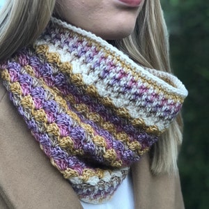 Può includere: Un scaldacollo all'uncinetto con un motivo a righe in tonalità di viola, marrone e bianco.