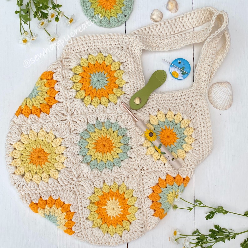 Puede incluir: Un bolso de mano de crochet con un dise&ntilde;o de girasol en tonos amarillos, verdes y crema. El bolso tiene una sola correa y est&aacute; hecho con un punto de granny square.