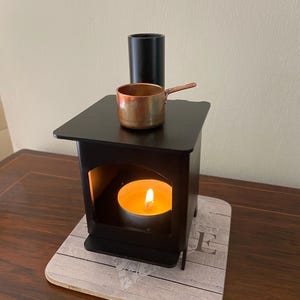 Pode incluir: Um porta-velas de metal preto em forma de fogão em miniatura com uma panela de cobre no topo. Uma vela pequena está acesa dentro do fogão.