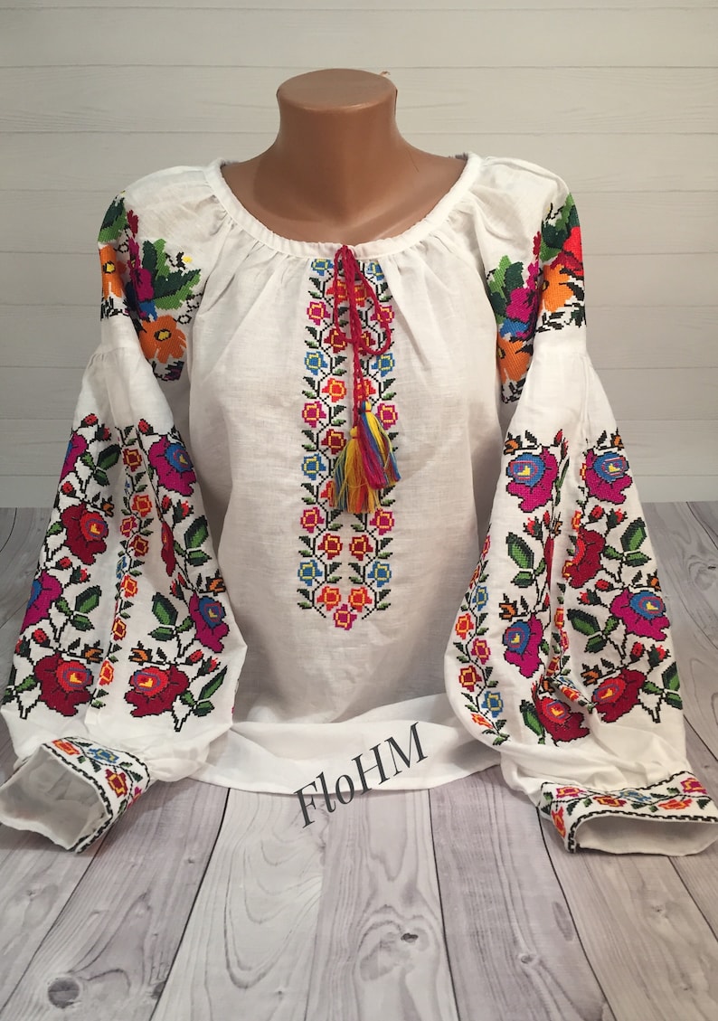 May include: White linen blouse with colorful embroidered floral patterns. The sleeves and neckline are trimmed with embroidery. The blouse has a red tassel.