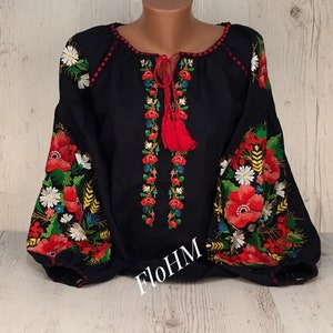 May include: A black embroidered blouse with red, white, and green floral designs. The blouse has a round neckline and long sleeves. The embroidery is detailed and intricate. The blouse is on a white wooden background.