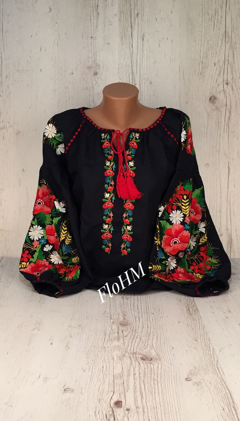 May include: A black embroidered blouse with red, white, and green floral designs. The blouse has a round neckline and long sleeves. The embroidery is detailed and intricate. The blouse is on a white wooden background.