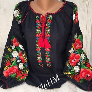 May include: A black linen blouse with red, white, and green embroidered flowers. The blouse has a round neckline and long sleeves. The embroidery is detailed and intricate. The text "FloHM" is visible on the front of the blouse.