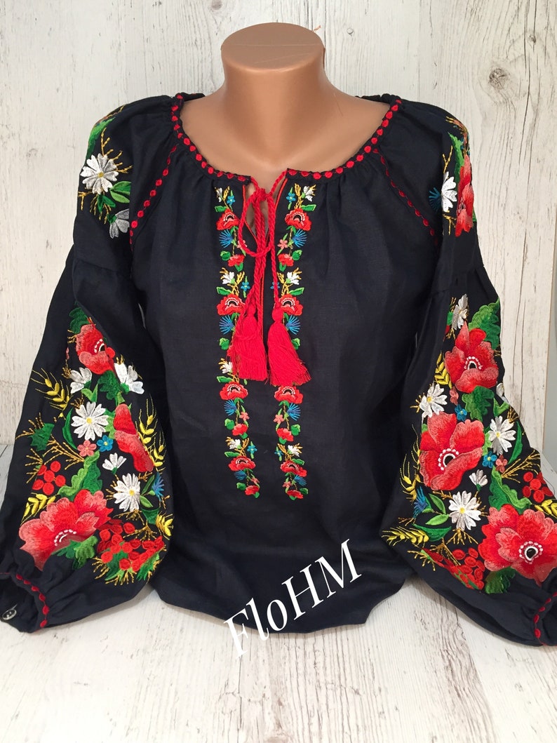 May include: A black linen blouse with red, white, and green embroidered flowers. The blouse has a round neckline and long sleeves. The embroidery is detailed and intricate. The text "FloHM" is visible on the front of the blouse.