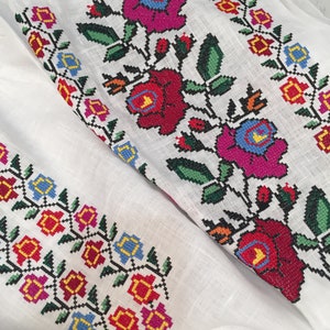 May include: White linen fabric with embroidered floral designs in red, pink, blue, yellow, and green. The flowers are stylized and have a traditional folk art look.