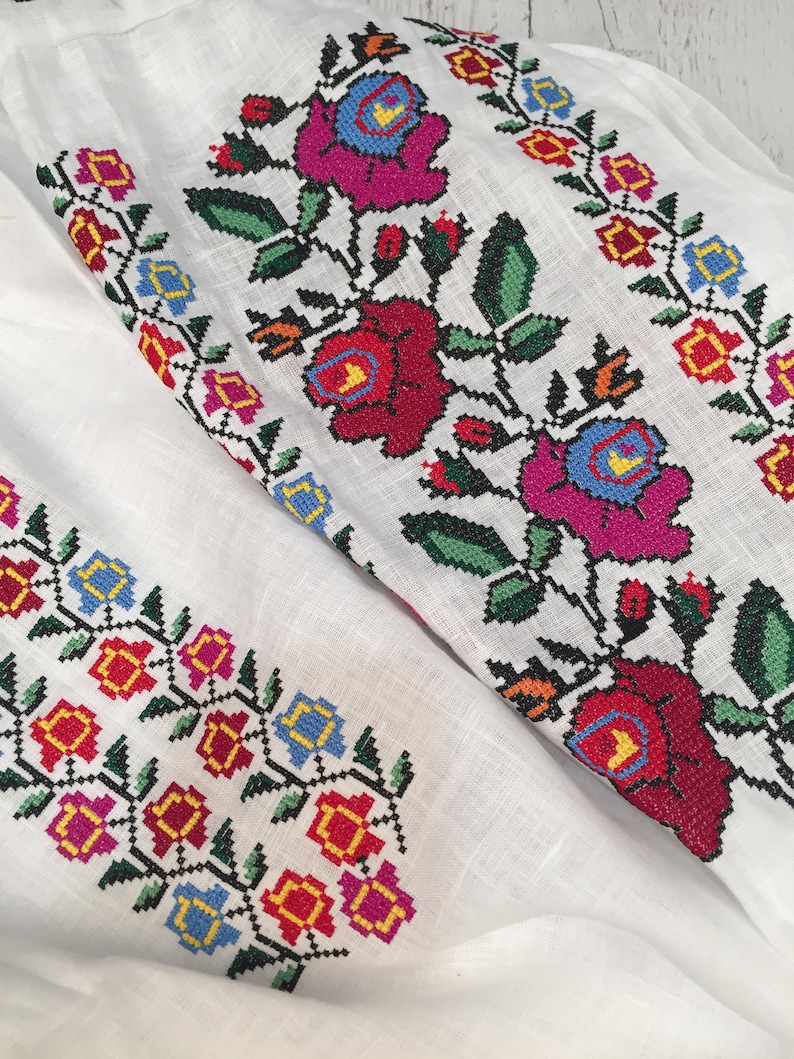 May include: White linen fabric with embroidered floral designs in red, pink, blue, yellow, and green. The flowers are stylized and have a traditional folk art look.