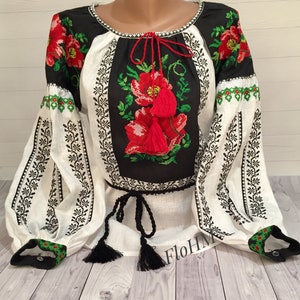 May include: A white linen blouse with black embroidery featuring red flowers and green leaves. The blouse has a black tie at the neckline and black tassels on the sleeves.
