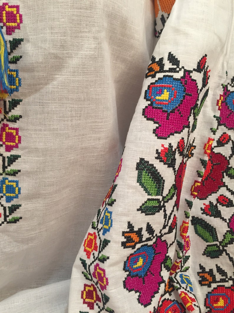 May include: White linen fabric with colorful embroidered floral designs. The embroidery features pink, blue, red, yellow, and green flowers with black outlines.